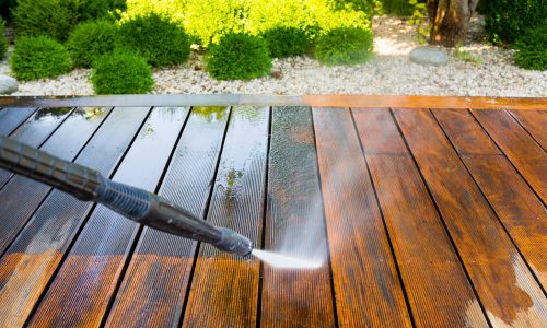 Power washing wooden deck surface, preparing for deck staining, surrounded by greenery and gravel.