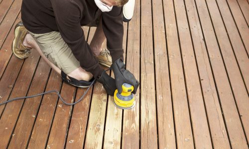Person using a power sander on a wooden deck, preparing the surface for staining as part of professional deck maintenance services by Valentine Property Services.